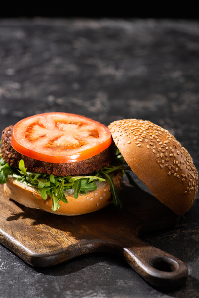 selective focus of tasty vegan burger with tomato and greens served on wooden board on textured surface - Photo, image