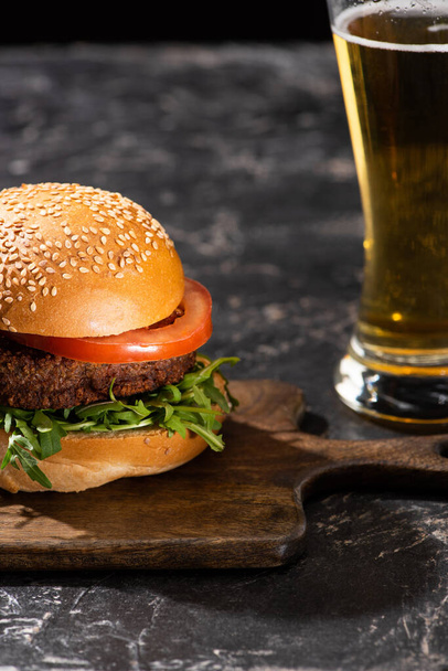 selective focus of tasty vegan burger with tomato and greens served on wooden board on textured surface with beer - Photo, Image