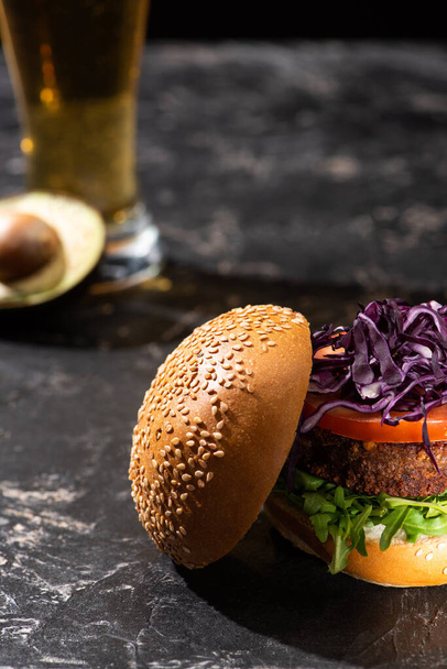 selective focus of tasty vegan burger with tomato, red cabbage and greens served on textured surface with beer and avocado - Photo, Image