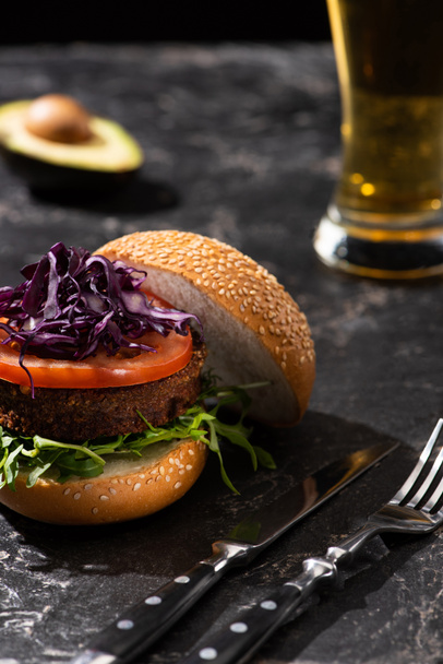 selective focus of tasty vegan burger with tomato, red cabbage and greens served on textured surface with cutlery, beer and avocado - Foto, Bild