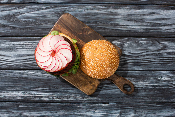 top view of tasty vegan burger with microgreens, radish, tomato on cutting board served on wooden table - Photo, Image