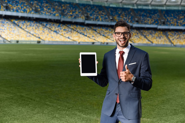 glücklicher junger Geschäftsmann in Anzug und Brille hält digitales Tablet mit leerem Bildschirm in der Hand und zeigt Daumen hoch im Stadion - Foto, Bild