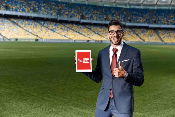 KYIV, UKRAINE - 20. června 2019: šťastný mladý podnikatel v obleku a brýlích, držící digitální tablet s aplikací Youtube a ukazující palec nahoru na stadionu - Fotografie, Obrázek