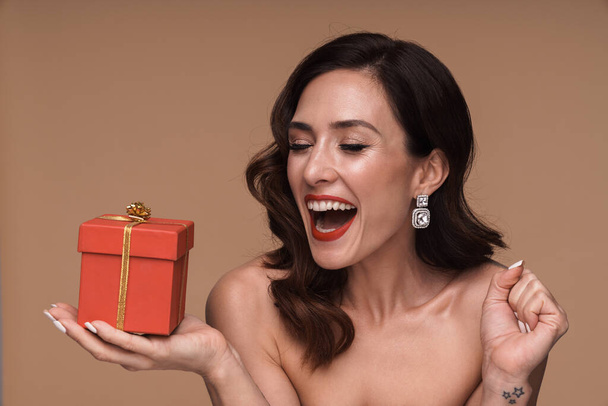Beauty portrait of happy gorgeous adult half-naked woman holding present box isolated over beige background - 写真・画像