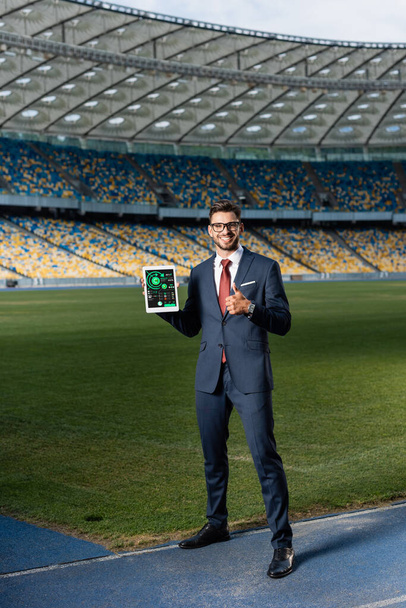 visão completa do jovem empresário feliz em terno e óculos segurando tablet digital com gráficos e gráficos na tela e mostrando polegar para cima no estádio
 - Foto, Imagem