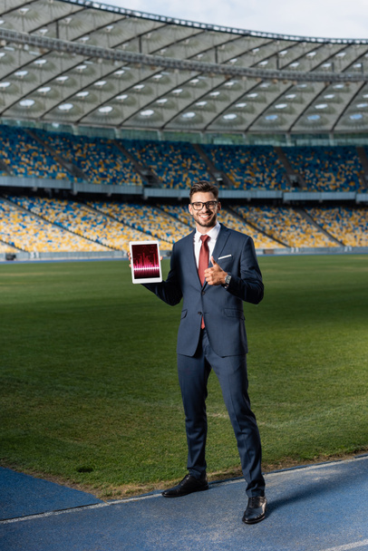 pełny widok długości szczęśliwy młody biznesmen w garniturze i okulary trzymając cyfrowy tablet z aplikacji handlowej i pokazując kciuk w górę na stadionie - Zdjęcie, obraz