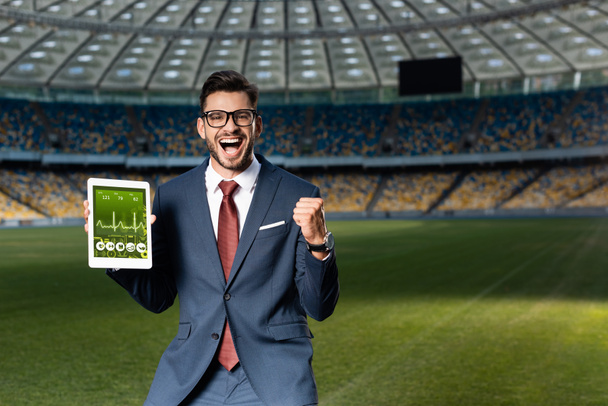 fröhlicher junger Geschäftsmann in Anzug und Brille hält digitales Tablet mit Gesundheits-App in der Hand und zeigt Ja-Geste im Stadion - Foto, Bild