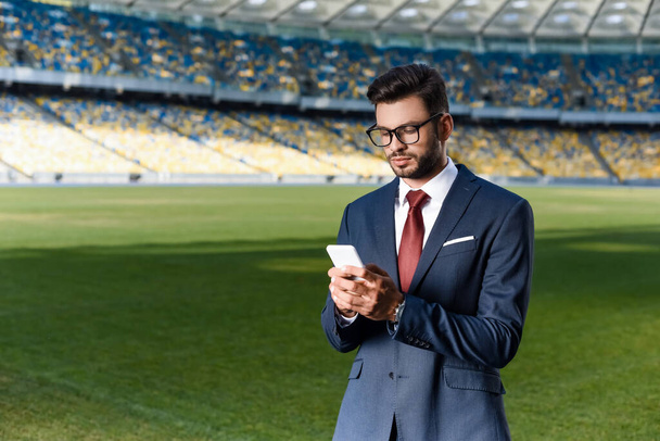 νεαρός επιχειρηματίας με κοστούμι και γυαλιά χρησιμοποιώντας smartphone στο γήπεδο - Φωτογραφία, εικόνα