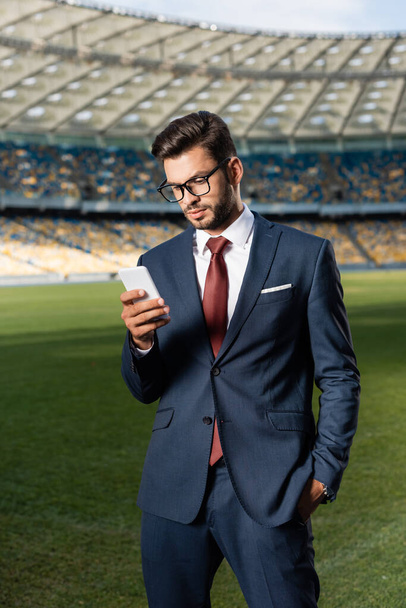 νεαρός επιχειρηματίας με κοστούμι και γυαλιά χρησιμοποιώντας smartphone στο γήπεδο - Φωτογραφία, εικόνα