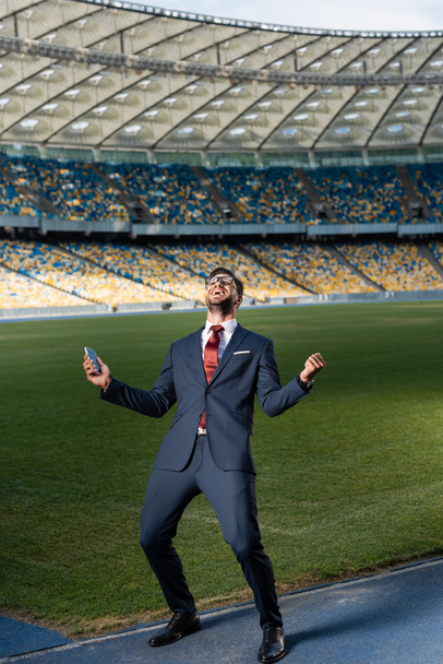 young businessman in suit and glasses holding smartphone and showing yeas gesture at stadium, sports betting concept - Zdjęcie, obraz