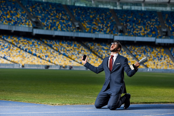 молодой бизнесмен в костюме, стоящий на коленях со смартфоном и трофеем на стадионе, концепция ставок на спорт
 - Фото, изображение