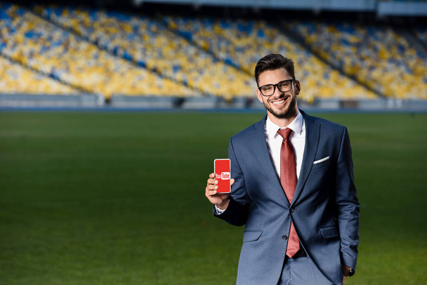 KYIV, UKRAINE - JUNE 20, 2019: smiling young businessman in suit and glasses holding smartphone with youtube app at stadium - Photo, image