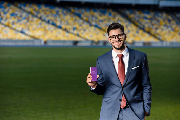 KYIV, UKRAINE - czerwiec 20, 2019: uśmiechnięty młody biznesmen w garniturze i okularach trzymający smartfona z aplikacją Instagram na stadionie - Zdjęcie, obraz
