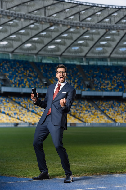 teljes hosszúságú kilátás izgatott fiatal üzletember öltöny és szemüveg gazdaság okostelefon üres képernyőn, és azt mutatja, igen gesztus a stadionban - Fotó, kép