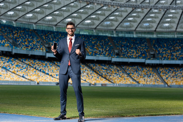 Lächelnder junger Geschäftsmann in Anzug und Brille hält Smartphone mit leerem Bildschirm in der Hand und zeigt Ja-Geste im Stadion - Foto, Bild