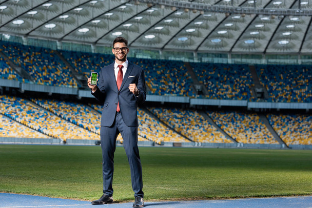 Lächelnder junger Geschäftsmann in Anzug und Brille hält Smartphone mit Gesundheits-App in der Hand und zeigt Ja-Geste im Stadion - Foto, Bild