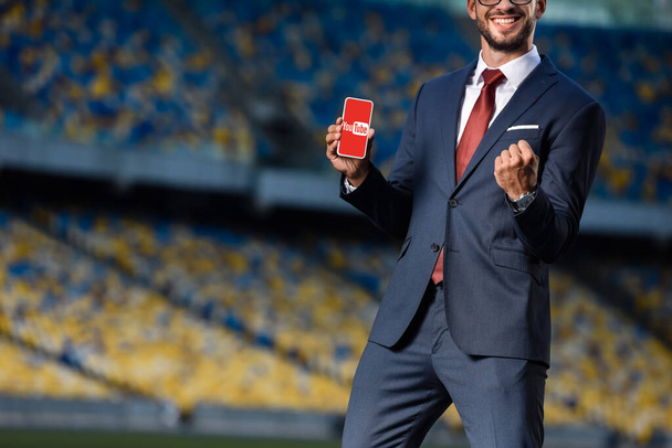 KYIV, UCRANIA - 20 de junio de 2019: vista recortada de un joven hombre de negocios sonriente con traje que sostiene el teléfono inteligente con la aplicación de youtube y muestra un gesto de sí en el estadio
 - Foto, Imagen