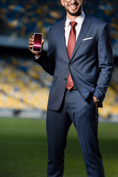 przycięty widok młodego biznesmena w garniturze z ręką w kieszeni gospodarstwa smartfon z aplikacją handlową na stadionie - Zdjęcie, obraz