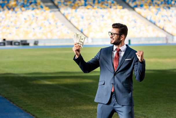 Улыбающийся молодой бизнесмен в костюме и очках с деньгами, показывающими жест "Да" на стадионе
 - Фото, изображение