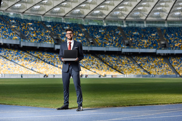 glimlachende jonge zakenman in pak met laptop in het stadion - Foto, afbeelding