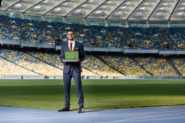 jeune homme d'affaires souriant en costume montrant ordinateur portable avec formation de football à l'écran au stade
 - Photo, image