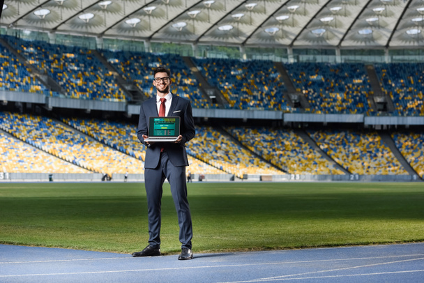 smiling young businessman in suit showing laptop with sports betting website at stadium - Photo, Image