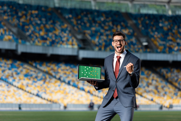 heureux jeune homme d'affaires en costume tenant ordinateur portable avec terrain de football et formation à l'écran et montrant geste oui au stade
 - Photo, image