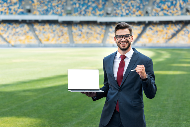 Lächelnder junger Geschäftsmann im Anzug zeigt Ja-Geste, während er Laptop mit leerem Bildschirm im Stadion hält - Foto, Bild