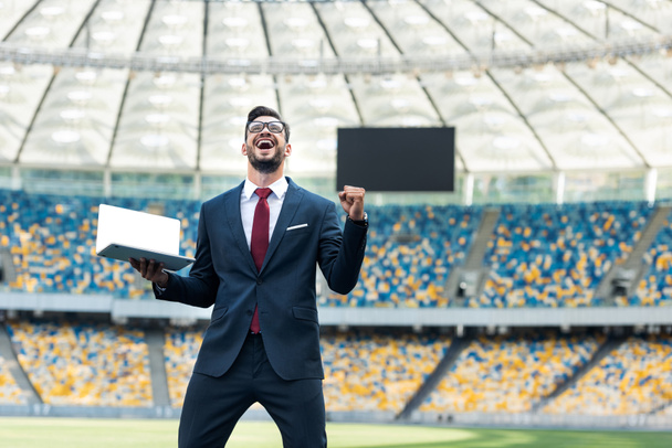 nízký úhel pohledu na šťastný mladý podnikatel v obleku ukazující ano gesto a držení notebook s prázdnou obrazovkou na stadionu - Fotografie, Obrázek