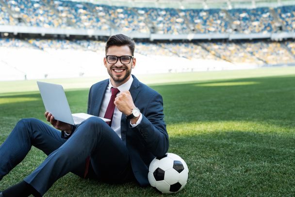 χαμογελαστός νεαρός επιχειρηματίας με κοστούμι με φορητό υπολογιστή και μπάλα ποδοσφαίρου κάθεται στο γήπεδο ποδοσφαίρου και δείχνει ναι χειρονομία στο γήπεδο, έννοια αθλητικών στοιχημάτων - Φωτογραφία, εικόνα