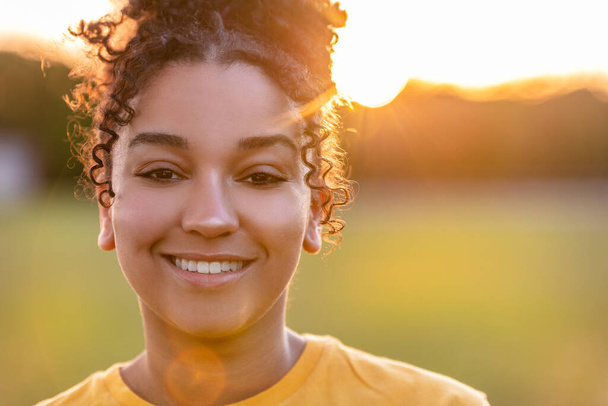 Beautiful biracial mixed race African American teenager teen girl young woman smiling with perfect teeth outside backlit at sunset or sunrise - Photo, Image