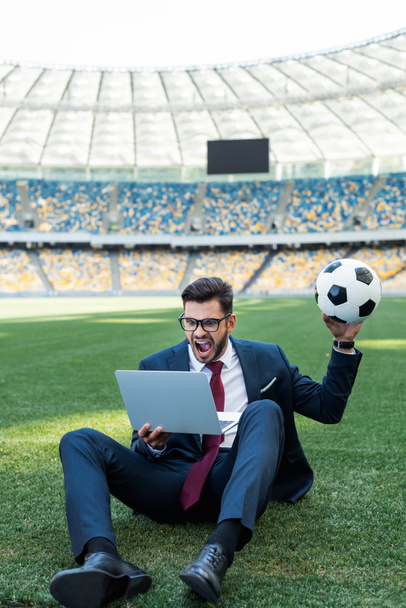 young businessman in suit with laptop and soccer ball sitting on football pitch at stadium and yelling, sports betting concept - Фото, изображение