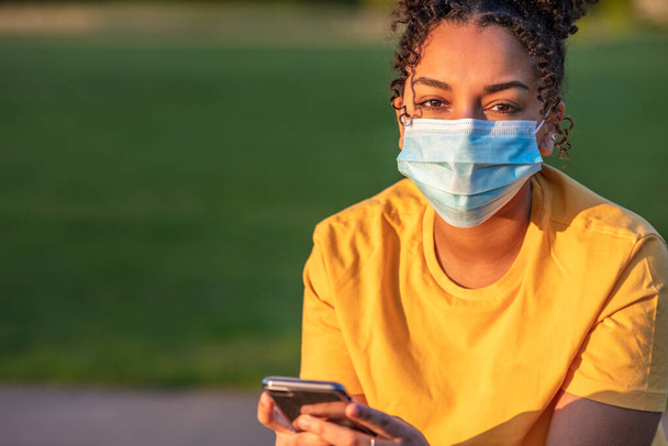 Mixed race biracial African American teenager teen girl young woman wearing a face mask and using mobile cell phone outside during the Coronavirus COVID-19 pandemic - Φωτογραφία, εικόνα