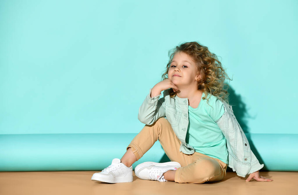 Cute little blond girl in casual clothing sits on floor hugging knee, tilting her head and smiling. Studio portrait on turquoise background - Foto, imagen