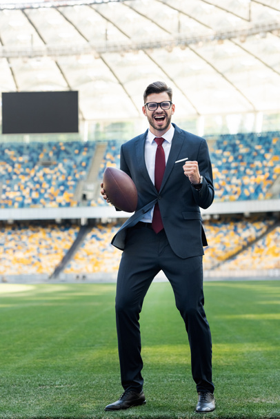 šťastný mladý podnikatel v obleku a brýle s rugby míč ukazuje ano gesto na stadionu - Fotografie, Obrázek