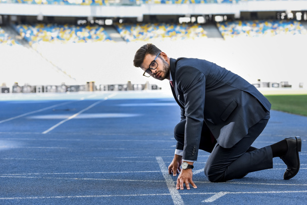 side view of young businessman in suit in start position on running track at stadium - Foto, imagen