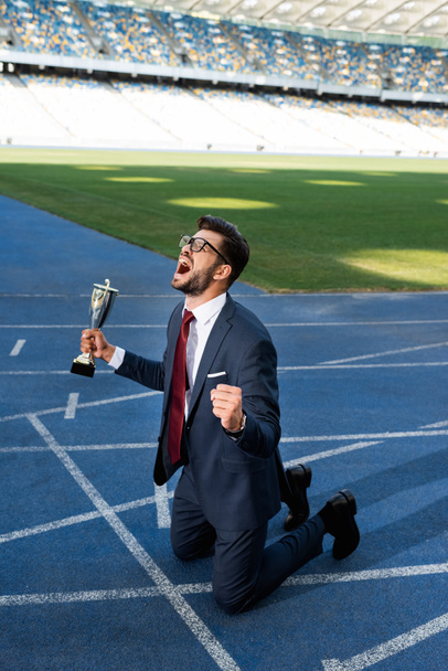 young businessman in suit standing on knees on running track with trophy and scream at stadium - Photo, Image