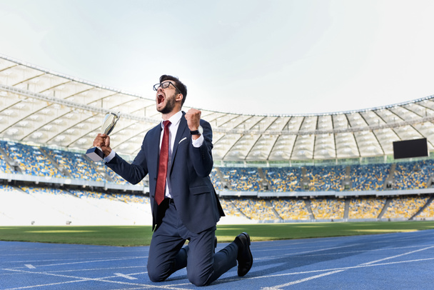 joven hombre de negocios en traje de pie de rodillas en la pista de atletismo con trofeo y gritar en el estadio
 - Foto, Imagen