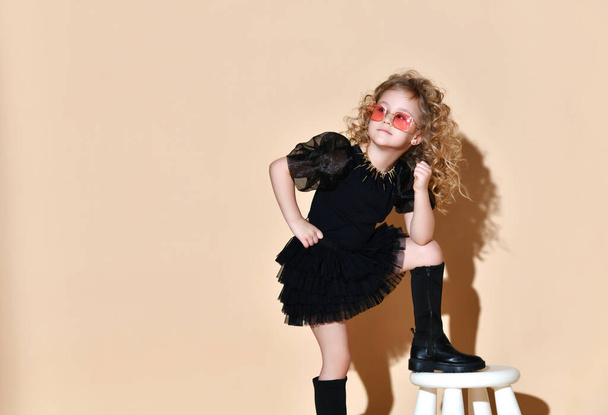 Blonde curly kid in orange sunglasses, necklace, black dress and boots. She put her foot on tabouret, posing on beige background - Photo, image