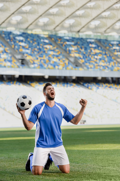 συναισθηματική επαγγελματίας ποδοσφαιριστής σε μπλε και άσπρο στολή με μπάλα στέκεται στα γόνατα στο γήπεδο ποδοσφαίρου και δείχνει ναι χειρονομία στο γήπεδο - Φωτογραφία, εικόνα
