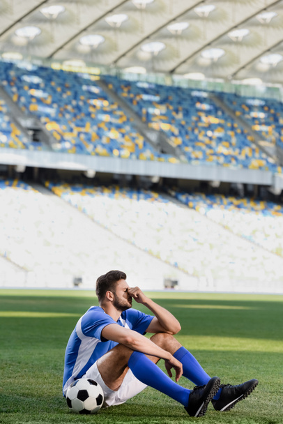 smutný profesionální fotbalista v modré a bílé uniformě sedí s míčem na fotbalovém hřišti na stadionu - Fotografie, Obrázek