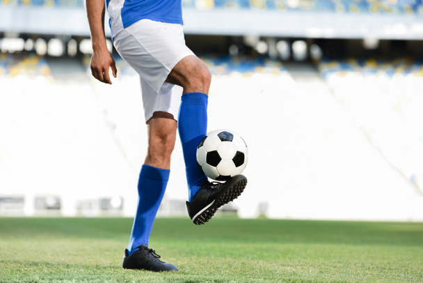 oříznutý pohled profesionálního fotbalisty v modré a bílé uniformě hrající s míčem na fotbalovém hřišti na stadionu - Fotografie, Obrázek