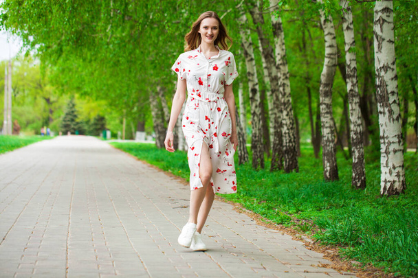 Full body portrait of a young beautiful blonde woman in white flowers dress walking spring street - Фото, изображение