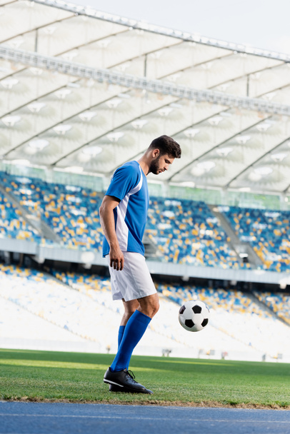zijaanzicht van professionele voetballer in blauw en wit uniform met bal op voetbalveld in het stadion - Foto, afbeelding