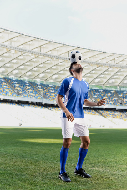 επαγγελματίας ποδοσφαιριστής σε μπλε και άσπρη στολή με μπάλα στο κεφάλι στο γήπεδο - Φωτογραφία, εικόνα