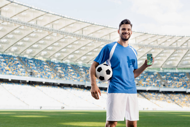 KYIV, UKRAINE - 20 ΙΟΥΝΙΟΥ 2019: χαμογελαστός επαγγελματίας ποδοσφαιριστής με μπλε και άσπρη στολή και μπάλα που δείχνει smartphone με εφαρμογές iphone στο γήπεδο - Φωτογραφία, εικόνα