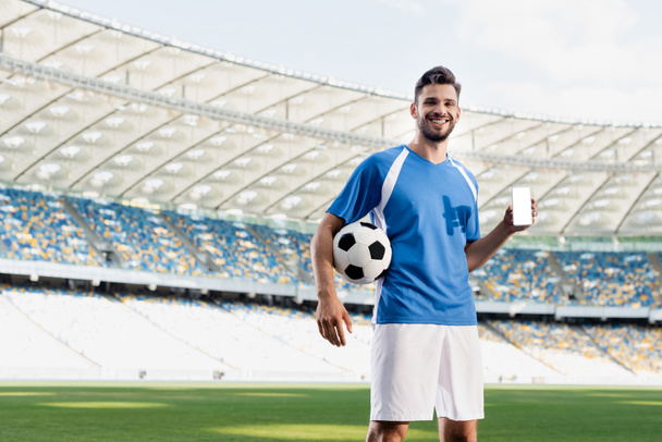 glimlachende professionele voetballer in blauw en wit uniform met bal tonen smartphone met blanco scherm in het stadion - Foto, afbeelding