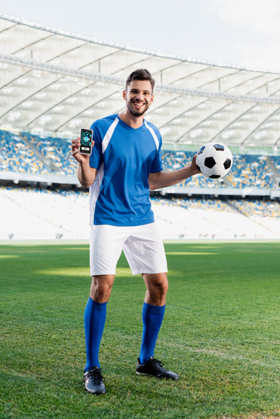 smiling professional soccer player in blue and white uniform with ball showing smartphone with stopwatch app at stadium - Photo, Image