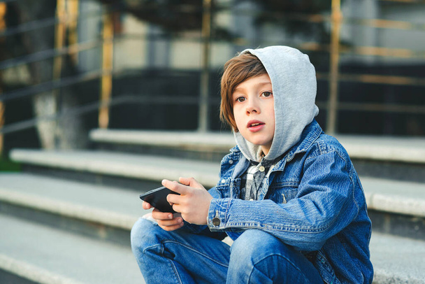 Schoolboy playing game on smartphone after school. Technology, lifestyle, leisure. Children addicted online games and videos. Boy use phone and plays games. Kid in the hood sitting on the stairs. - Photo, Image