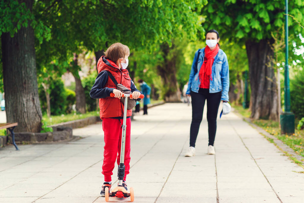 Family in safety masks outdoors. Riding boy on scooter in park. Boy wears medical face mask. Real life 2020. Coronavirus epidemic. Mother and son on a walk during coornavirus quarantine. - Photo, Image